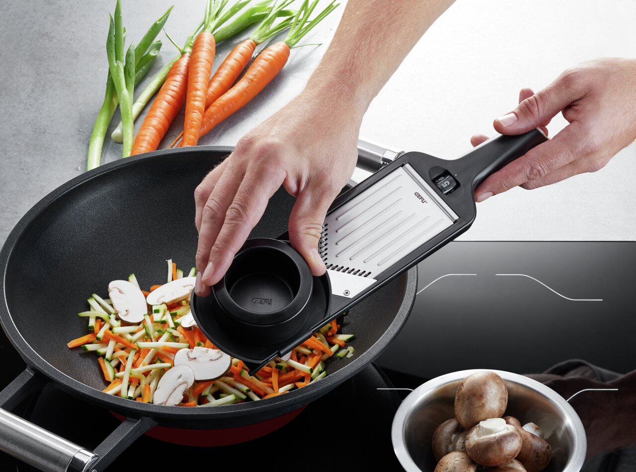 Bovenaanzicht: iemand gebruikt een Julienne VIOLI 2.0-schaaf om groenten in een pan te snijden. Op de achtergrond zijn wortelen, asperges en champignons te zien.