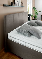 Seitliche Perspektive auf ein elegantes Polsterbett mit grauem Kopfteil und modernen Kissen in einem stilvoll eingerichteten Schlafzimmer.