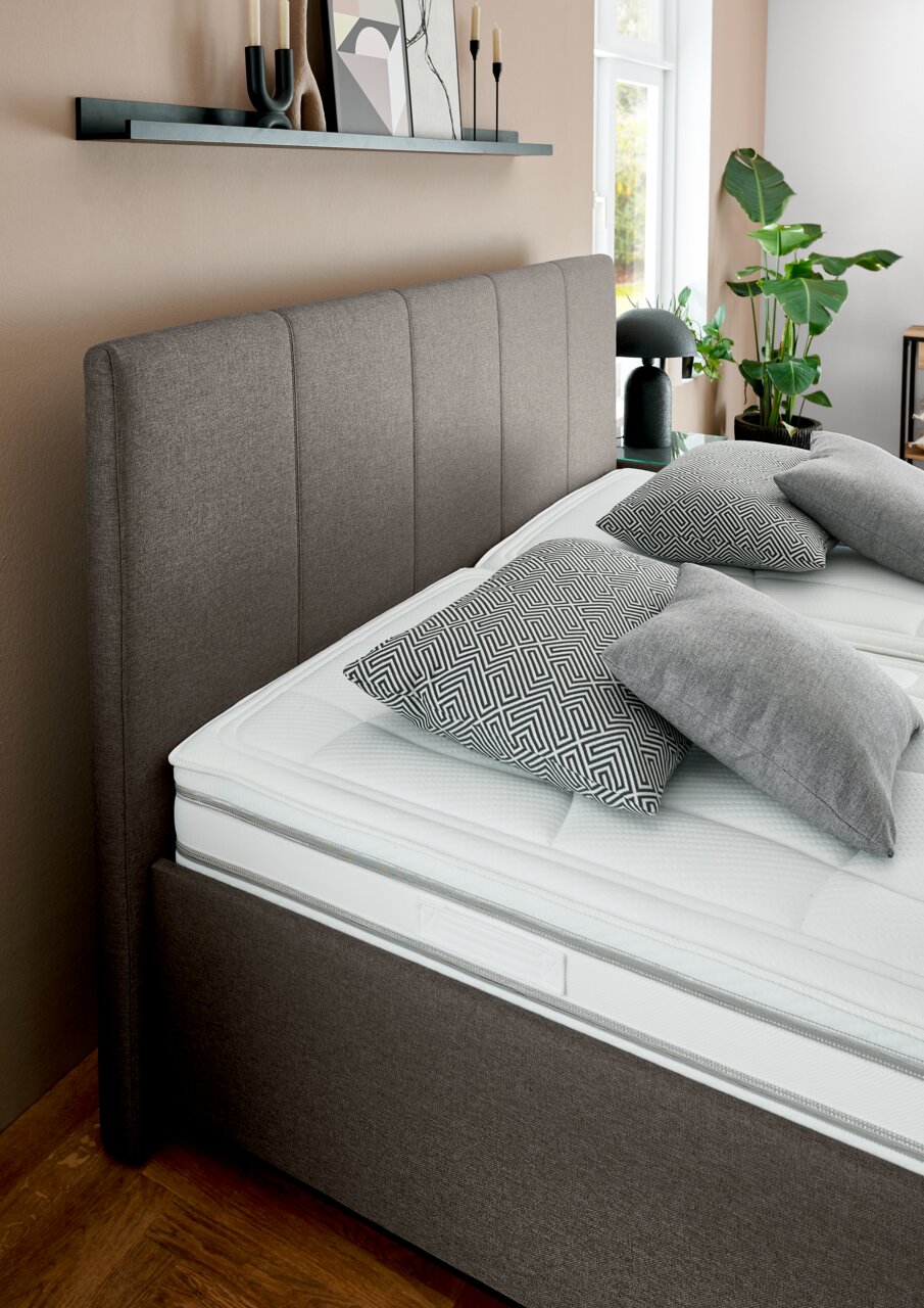 Seitliche Perspektive auf ein elegantes Polsterbett mit grauem Kopfteil und modernen Kissen in einem stilvoll eingerichteten Schlafzimmer.
