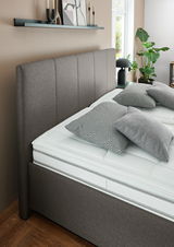 Seitliche Perspektive auf ein elegantes Polsterbett mit grauem Kopfteil und modernen Kissen in einem stilvoll eingerichteten Schlafzimmer.