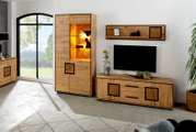 Moderne entertainmentset voor de woonkamer van licht massief hout, bestaande uit een hoge vitrinekast met warm verlichte glazen voorkant, een tv-lowboard en een wandplank, gezien vanaf de zijkant links.
