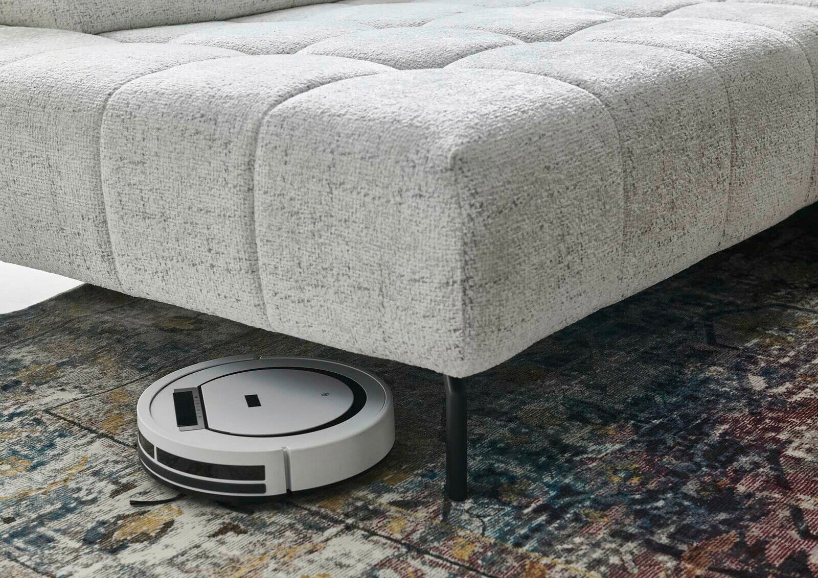 Eckgarnitur in hellem Stoffbezug mit einem Saugroboter, der darunter reinigt, aus seitlicher Perspektive.