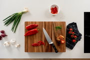Draufsicht auf ein Holzschneidebrett mit Küchenmesser, roten Paprika und geschnittenen Paprikastreifen, daneben Frühlingszwiebeln, Knoblauch, rote Zwiebeln, gehackte Kräuter, Cherrytomaten auf einem dunklen Küchentuch und eine Schale mit Paprikastreifen a
