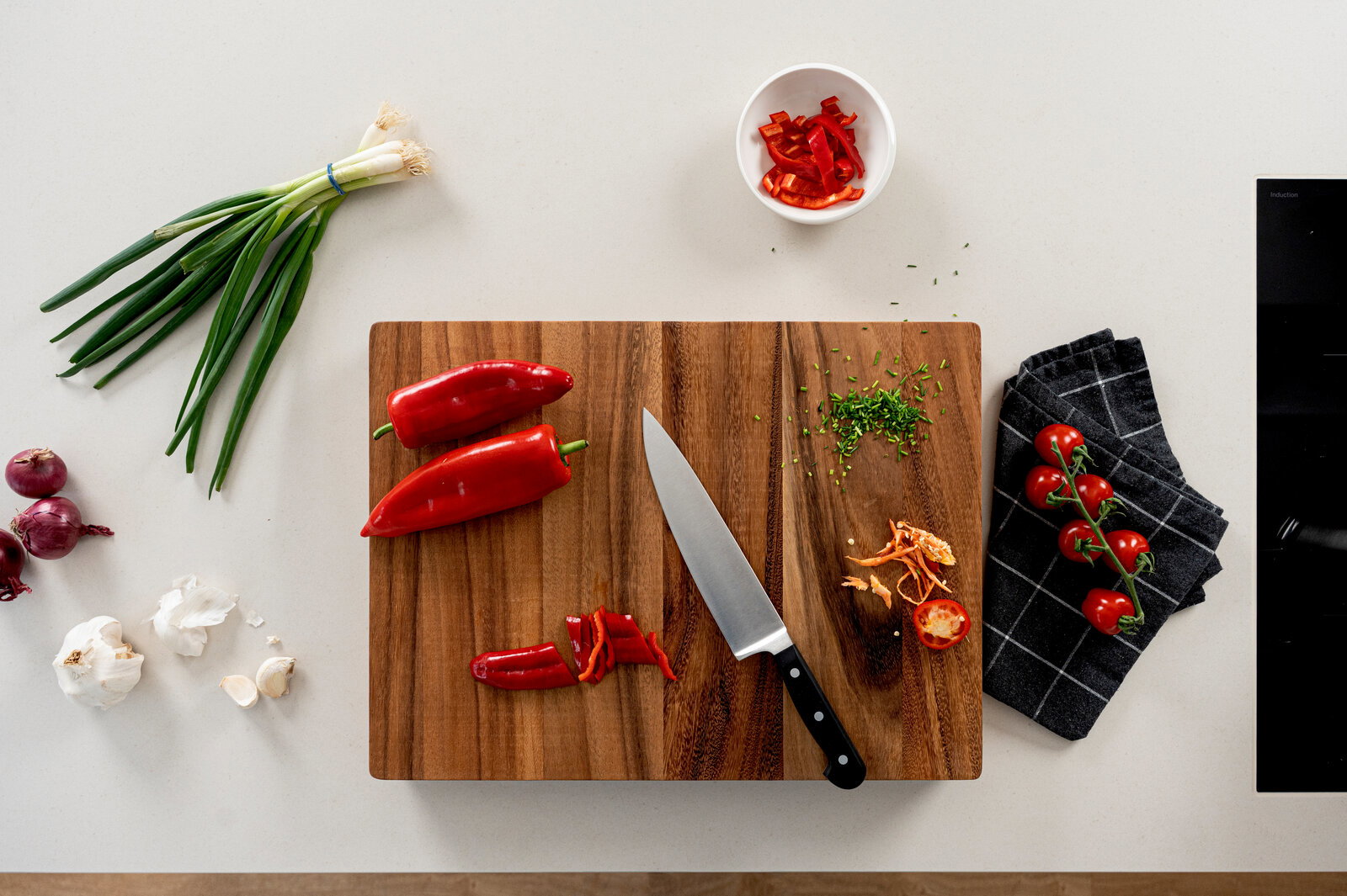 Draufsicht auf ein Holzschneidebrett mit Küchenmesser, roten Paprika und geschnittenen Paprikastreifen, daneben Frühlingszwiebeln, Knoblauch, rote Zwiebeln, gehackte Kräuter, Cherrytomaten auf einem dunklen Küchentuch und eine Schale mit Paprikastreifen a