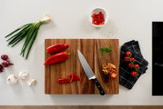 Draufsicht auf ein Holzschneidebrett mit Küchenmesser, roten Paprika und geschnittenen Paprikastreifen, daneben Frühlingszwiebeln, Knoblauch, rote Zwiebeln, gehackte Kräuter, Cherrytomaten auf einem dunklen Küchentuch und eine Schale mit Paprikastreifen a