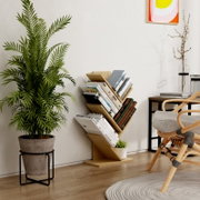 Schräg von vorn fotografiertes, modernes Bücherregal aus hellem Holz mit diagonal gestapelten Büchern in einem minimalistischen Wohnraum neben Zimmerpflanze und Sessel.