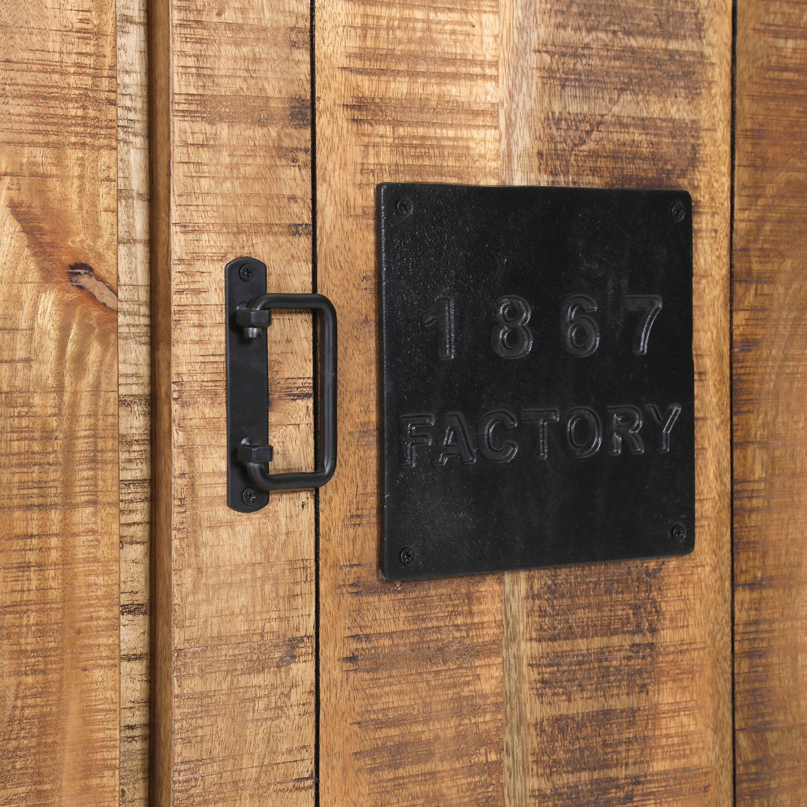 Nahaufnahme eines rustikalen Holzschranks mit schwarzem Metallgriff und einer Metallplatte mit der Aufschrift '1867 FACTORY'.