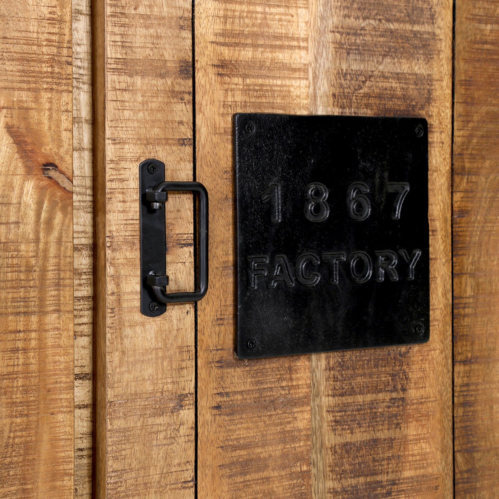 Close-up van een rustieke houten kast met een zwarte metalen handgreep en een metalen plaatje met het opschrift '1867 FACTORY'.