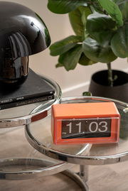 Orangefarbene Flip-Uhr auf einem gläsernen Beistelltisch, seitliche Perspektive.