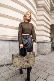 Frau mit einer großen, gemusterten Tasche in der Hand, die auf einem städtischen Gehweg vor einem Gebäude mit dekorativer Fassade geht. Die Tasche hat einen stabilen, schwarzen Griff. Perspektive: seitlich.