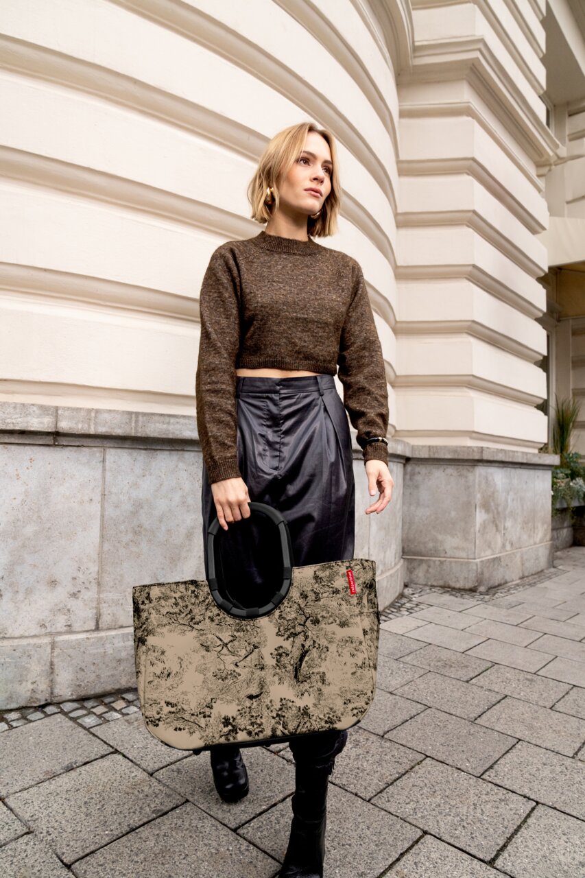 Frau mit einer großen, gemusterten Tasche in der Hand, die auf einem städtischen Gehweg vor einem Gebäude mit dekorativer Fassade geht. Die Tasche hat einen stabilen, schwarzen Griff. Perspektive: seitlich.