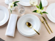 Elegant gedeckter Esstisch mit weißem Geschirr, goldfarbenem Besteck, gefalteter weißer Serviette, Wasser- und Weinglas sowie kleinen weißen Blumenvasen auf hellem Holztisch, Draufsicht von oben