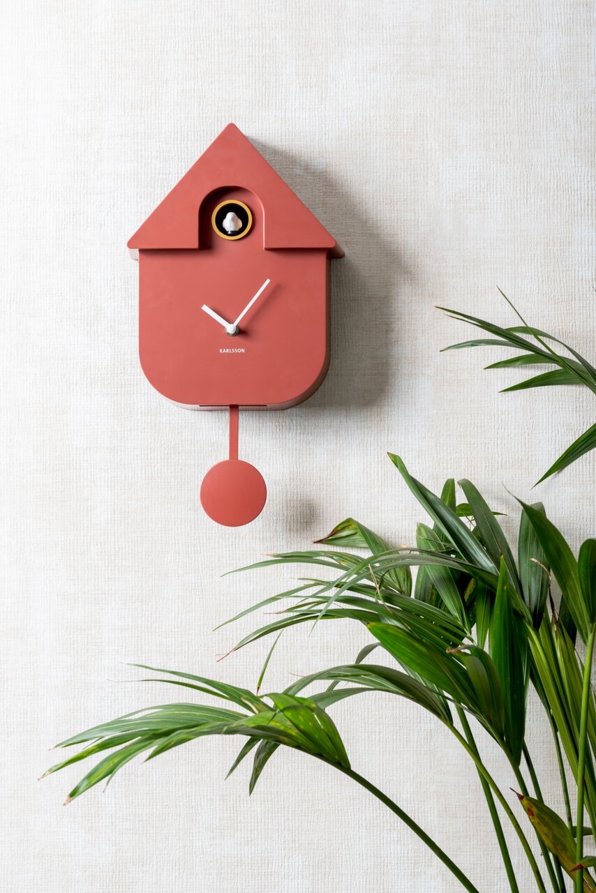 Moderne rote Kuckucksuhr von KARLSSON an einer Wand, mit einem Pendel und umgeben von grünen Pflanzen, seitliche Perspektive.