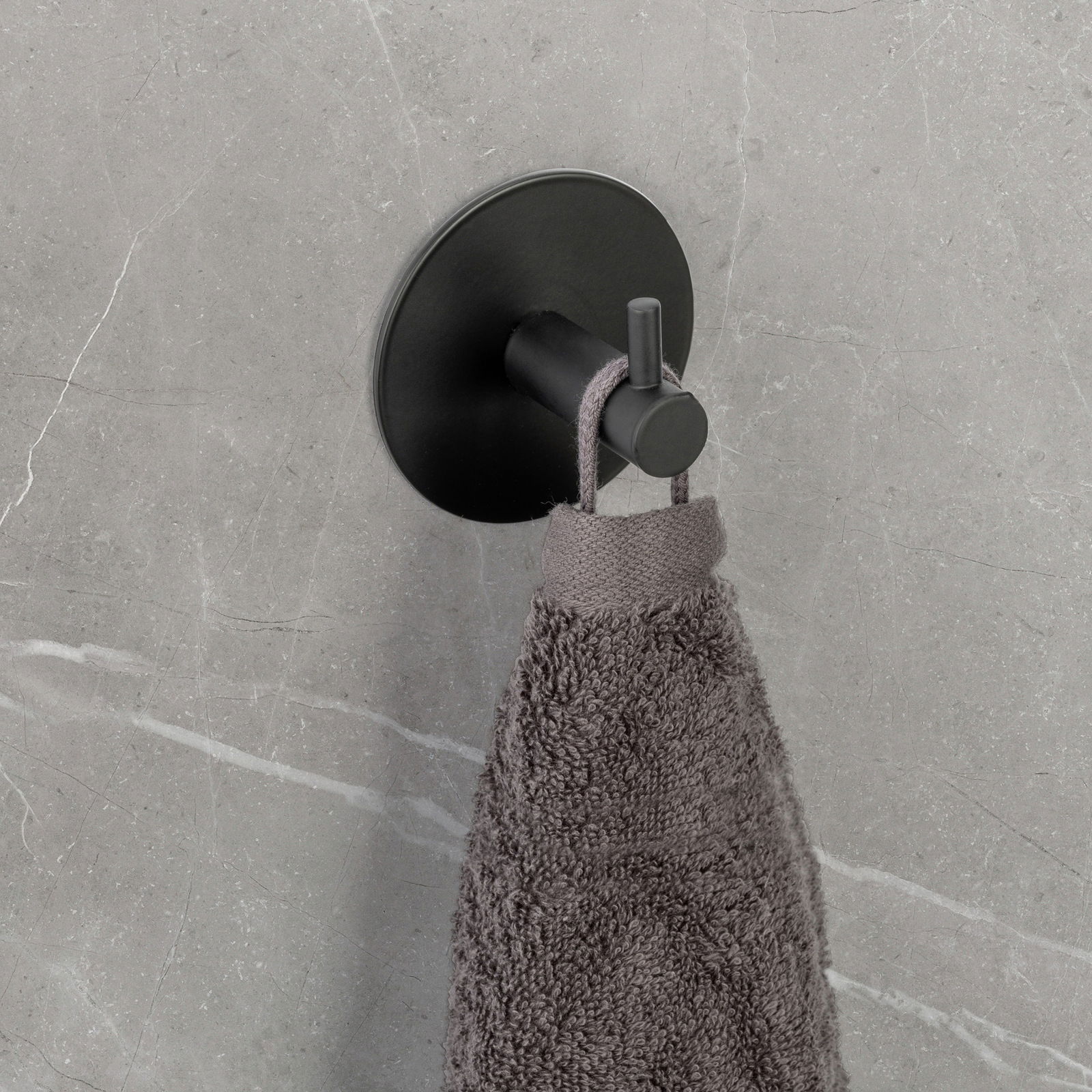 Schwarzer Wandhaken Uno Meleto an einer grauen Wand, von der Seite fotografiert, mit einem grauen Handtuch daran hängend.