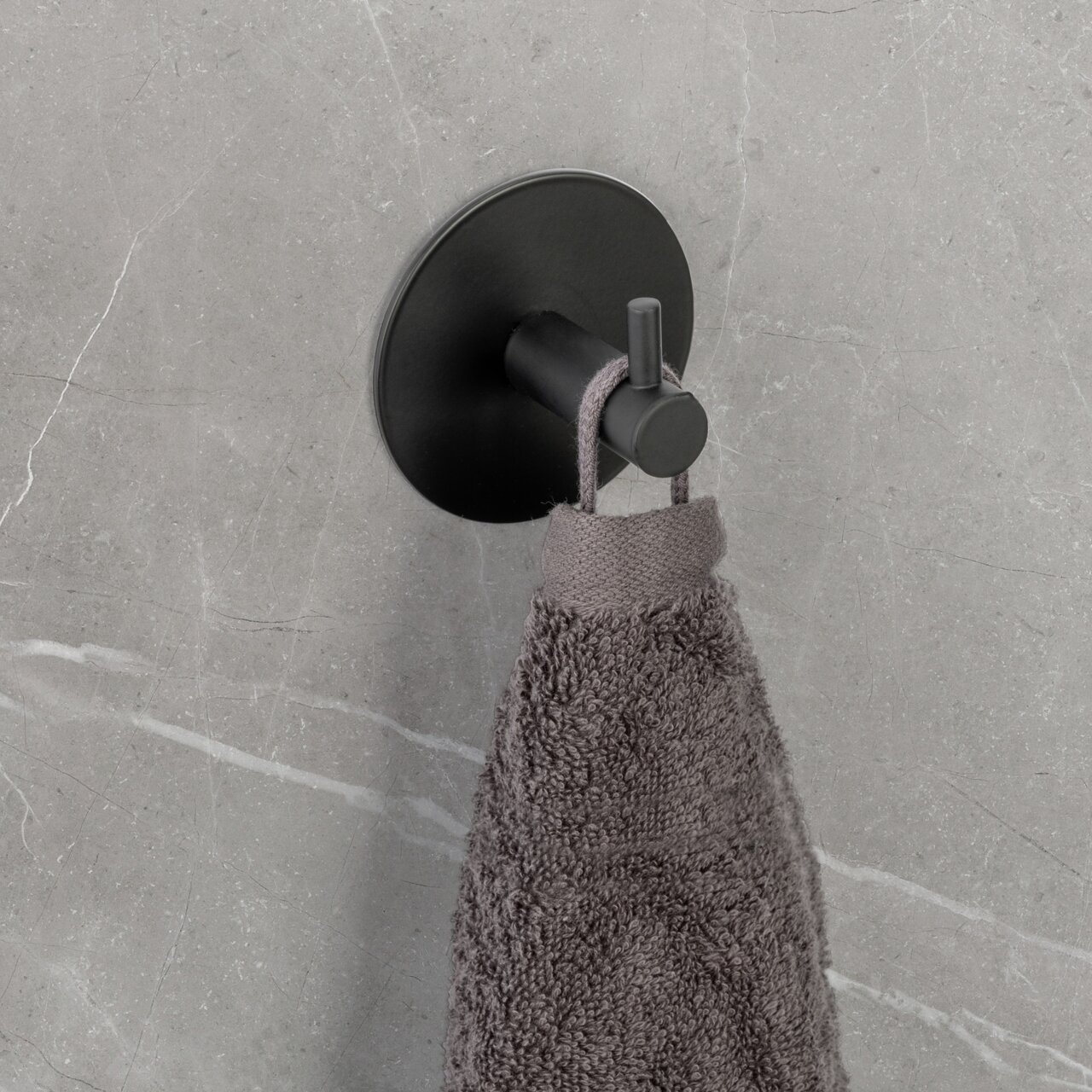Schwarzer Wandhaken Uno Meleto an einer grauen Wand, von der Seite fotografiert, mit einem grauen Handtuch daran hängend.
