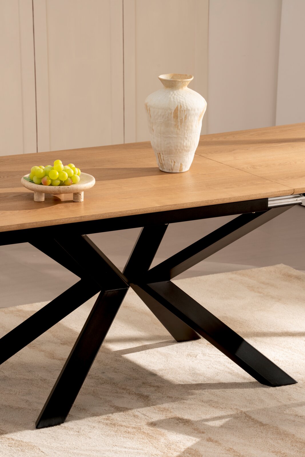 Moderner Esstisch mit warmer Holzplatte und schwarzem X-förmigem Metallgestell, darauf eine helle Keramikvase und eine Schale mit grünen Trauben, aufgenommen aus seitlicher Perspektive auf Augenhöhe