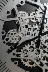 Nahaufnahme einer silberfarbenen Wanduhr aus Metall mit sichtbaren Zahnrädern und schwarzen Zeigern, von vorne betrachtet.