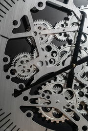 Nahaufnahme einer silberfarbenen Wanduhr aus Metall mit sichtbaren Zahnrädern und schwarzen Zeigern, von vorne betrachtet.