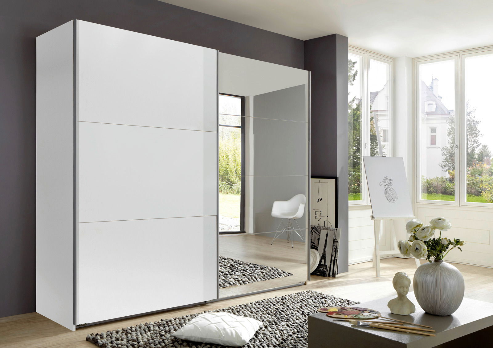 Moderner, zweiflügeliger Schwebetürenschrank in einem hellen Raum mit großen Fenstern, einer weißen Tür und einer Spiegeltür, seitliche Perspektive.