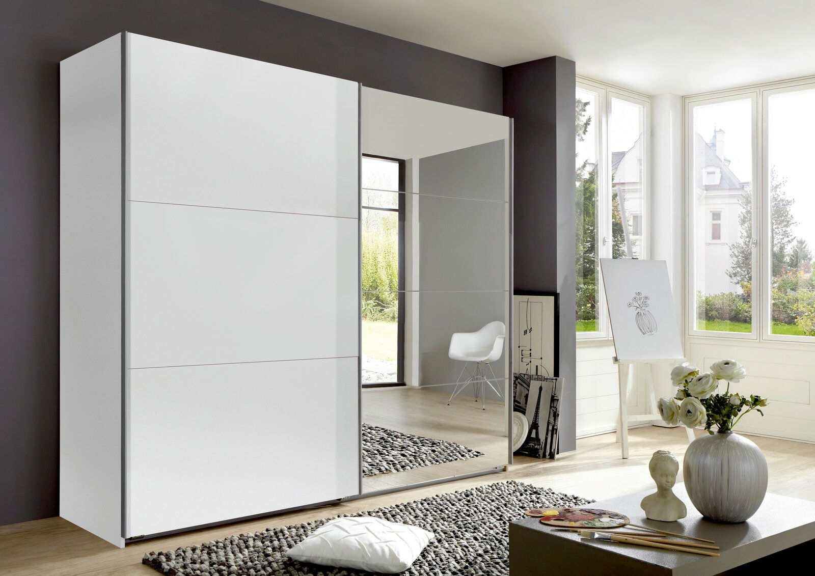 Moderner, zweiflügeliger Schwebetürenschrank in einem hellen Raum mit großen Fenstern, einer weißen Tür und einer Spiegeltür, seitliche Perspektive.