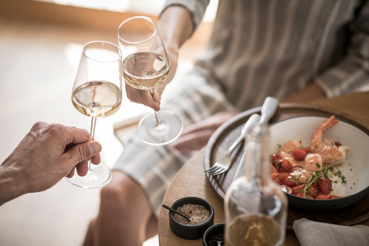 Twee mensen proosten met glazen Riesling, op de achtergrond een bord met garnalen en tomaten, gezien van bovenaf.