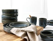 Verschiedene dunkelgraue Keramikteller und -schalen, gestapelt auf einem Holzbrett, mit zwei Tassen im Hintergrund. Perspektive von schräg oben.