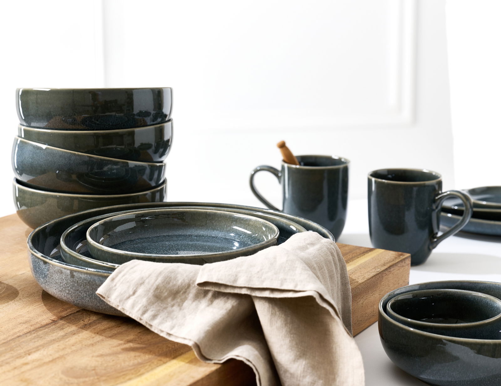 Verschiedene dunkelgraue Keramikteller und -schalen, gestapelt auf einem Holzbrett, mit zwei Tassen im Hintergrund. Perspektive von schräg oben.