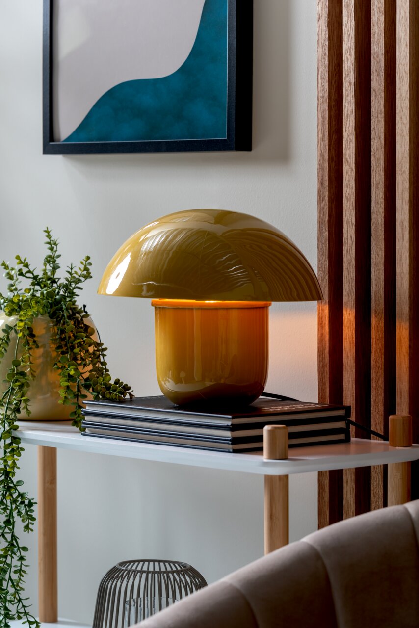 Leitmotiv Tischleuchte MUSHROOM Gelbe Tischleuchte mit gewölbtem Schirm auf einem Regal, umgeben von Pflanzen und Büchern, seitliche Perspektive.