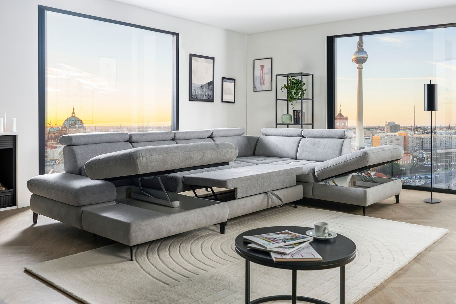 Graue Rundecke mit verstellbaren Kopfstützen und ausziehbarer Liegefläche in einem modernen Wohnzimmer mit Blick auf die Stadt. Perspektive von vorne links.