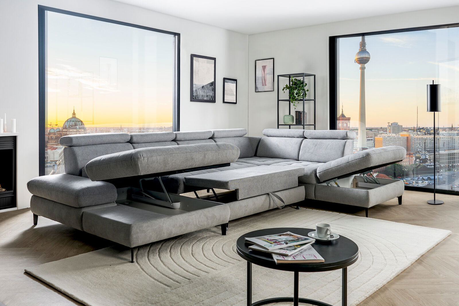 Graue Rundecke mit verstellbaren Kopfstützen und ausziehbarer Liegefläche in einem modernen Wohnzimmer mit Blick auf die Stadt. Perspektive von vorne links.
