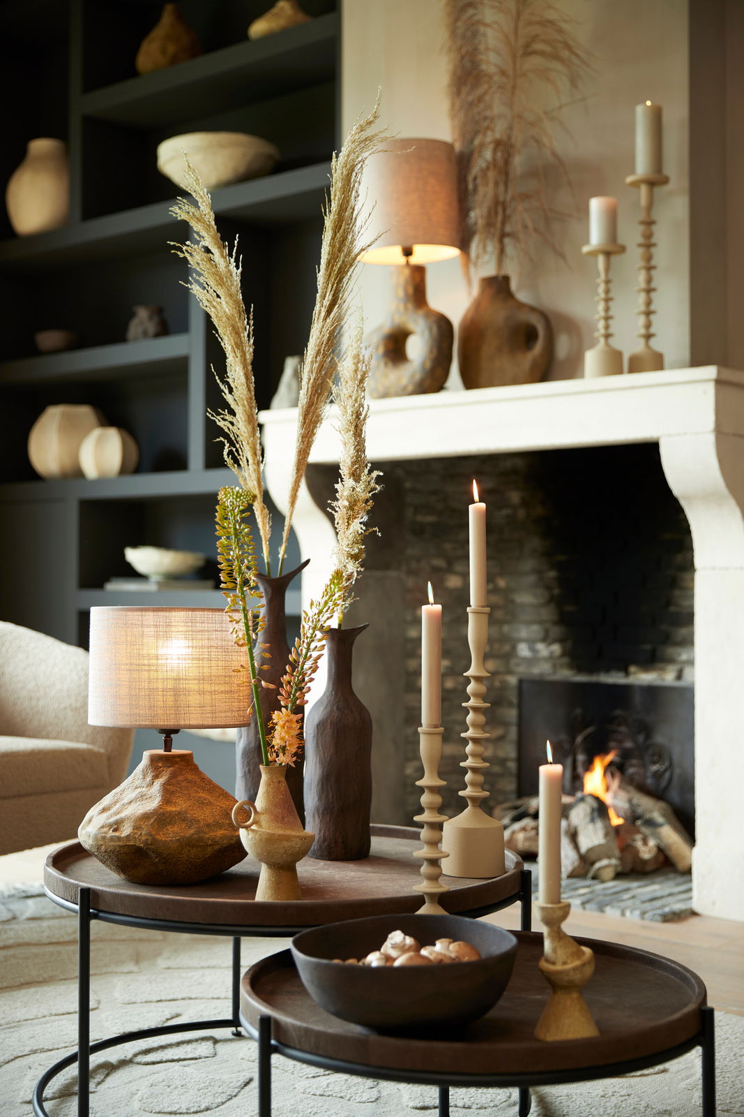 Gemütliches Wohnzimmer mit einem modernen Lampenschirm in Zylinderform auf einem Tisch, umgeben von Kerzen und dekorativen Vasen, im Hintergrund ein Kaminfeuer. Perspektive von vorne.