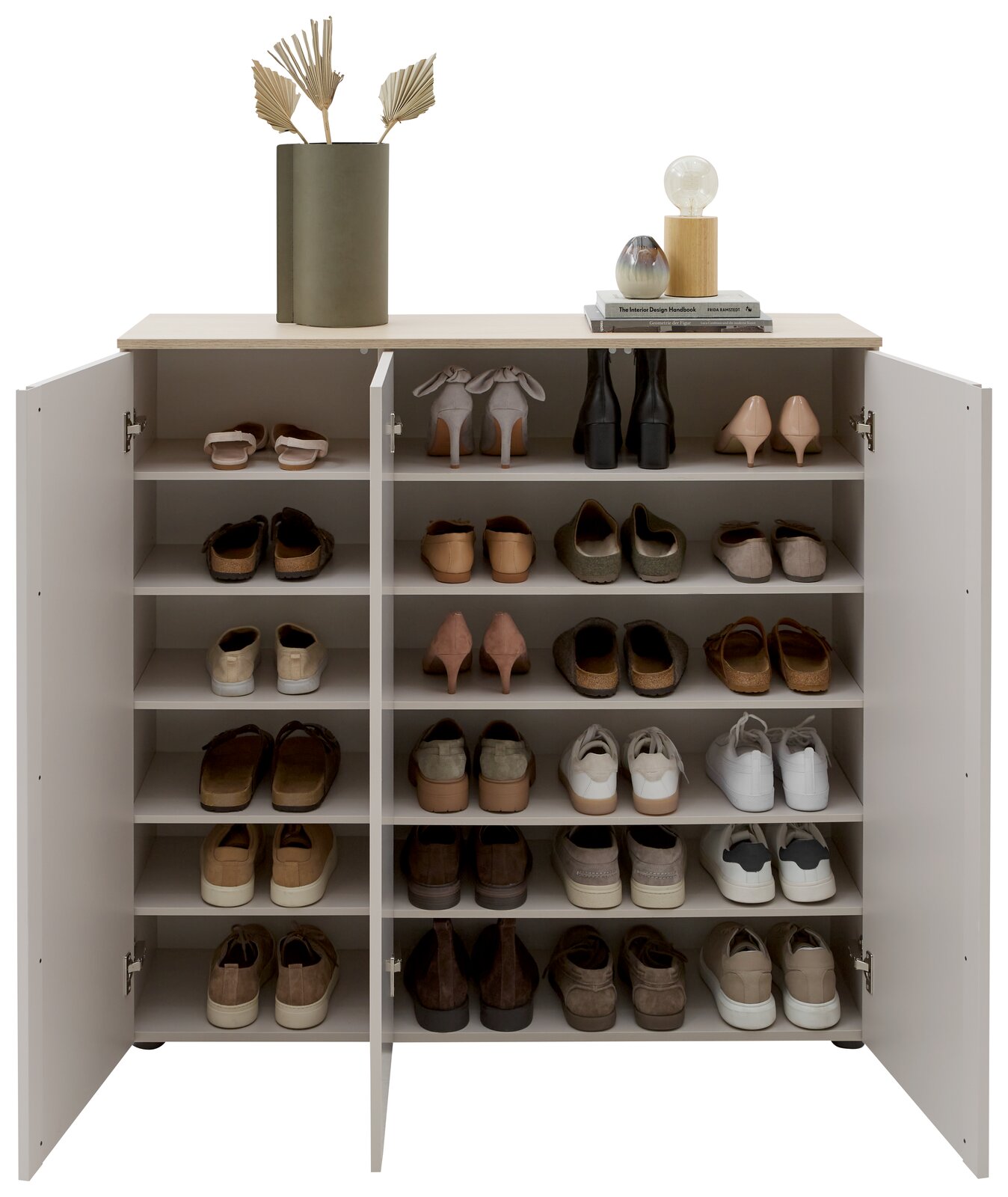 Offener weißer Schuhschrank mit mehreren Regalböden und drei Türen, gefüllt mit verschiedenen Schuhpaaren; Draufsicht leicht frontal von vorn, oben dekoriert mit Vase, Büchern und kleiner Lampe.