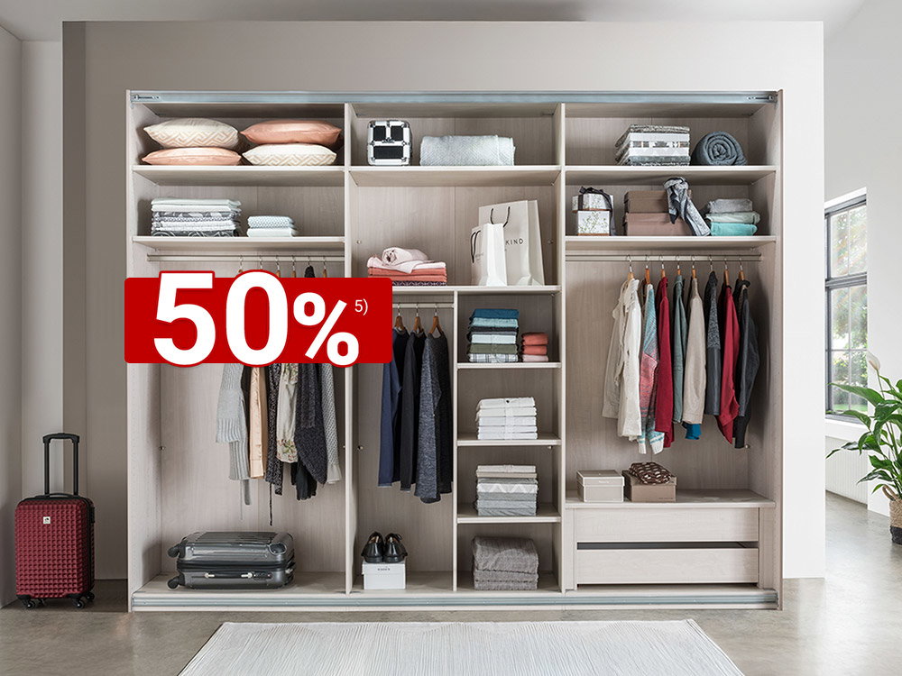 Großer offener Kleiderschrank mit Fächern, Kleiderstangen und Schubladen, ordentlich gefüllt mit Kleidung, Kissen und Boxen, daneben ein Koffer