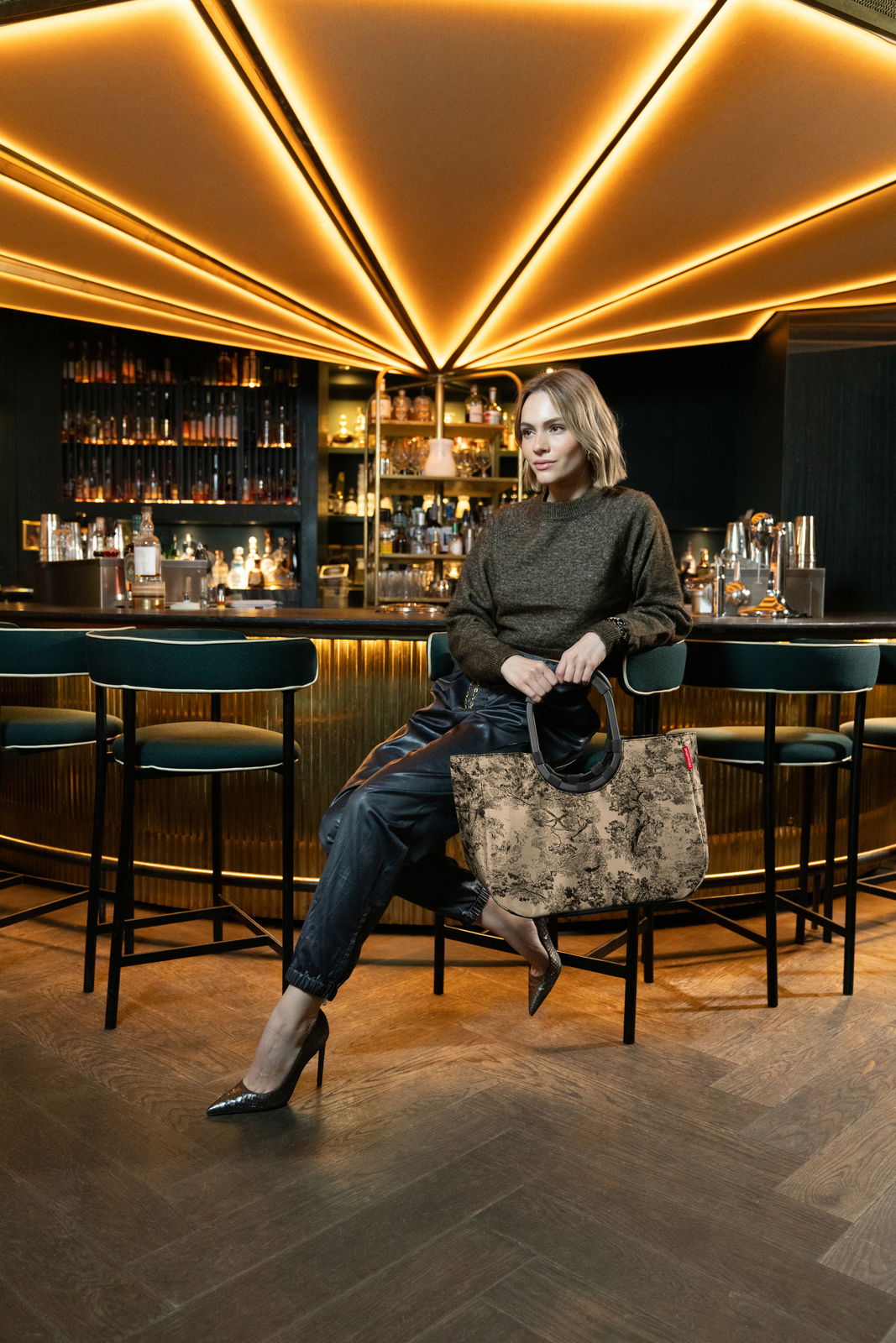Frau sitzt in einer stilvollen Bar und hält eine große, gemusterte Tasche mit runden Griffen. Die Perspektive zeigt die Tasche von vorne.