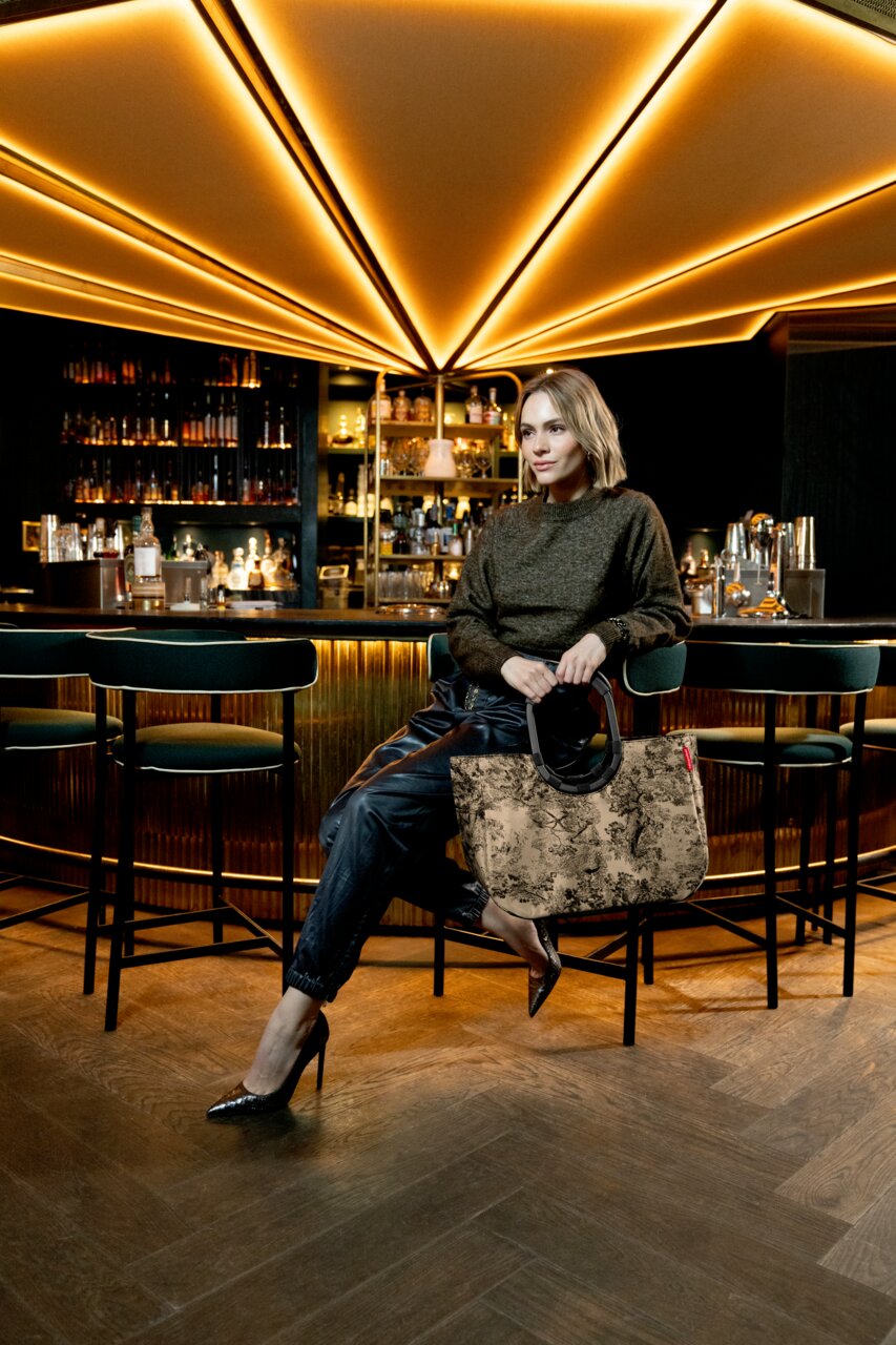 Frau sitzt in einer stilvollen Bar und hält eine große, gemusterte Tasche mit runden Griffen. Die Perspektive zeigt die Tasche von vorne.