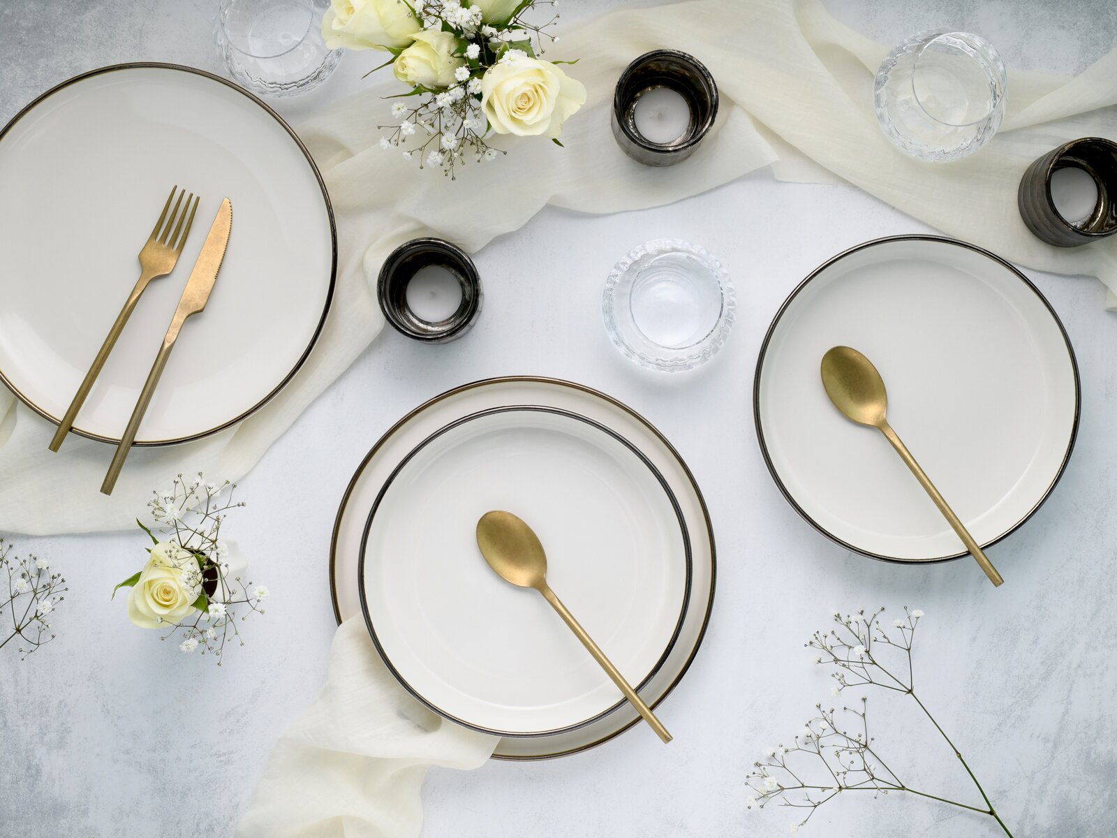 Draufsicht auf elegant gedeckten Tisch mit weißen Tellern mit dunklem Rand, goldfarbenem Besteck, Glas-Teelichthaltern, Wassergläsern und weißen Rosen auf hellem Stoff