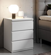 Weiße Nachtkommode mit drei Schubladen, darauf eine runde Lampe und ein Buch, seitliche Perspektive