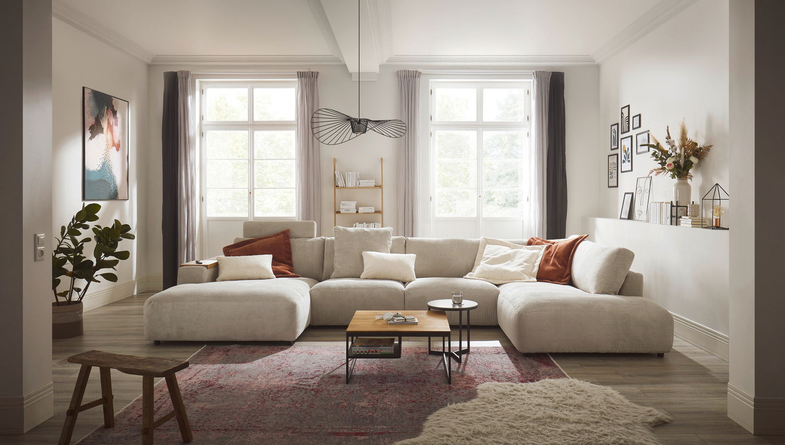 Musterring Ecksofa MR LUCIA Gemütliches Wohnzimmer mit großem, beigem Ecksofa Musterring LUCIA, dekoriert mit Kissen in Erdtönen, aus der Frontalperspektive fotografiert. Im Hintergrund sind große Fenster, ein Bücherregal und Wanddekorationen zu sehen.