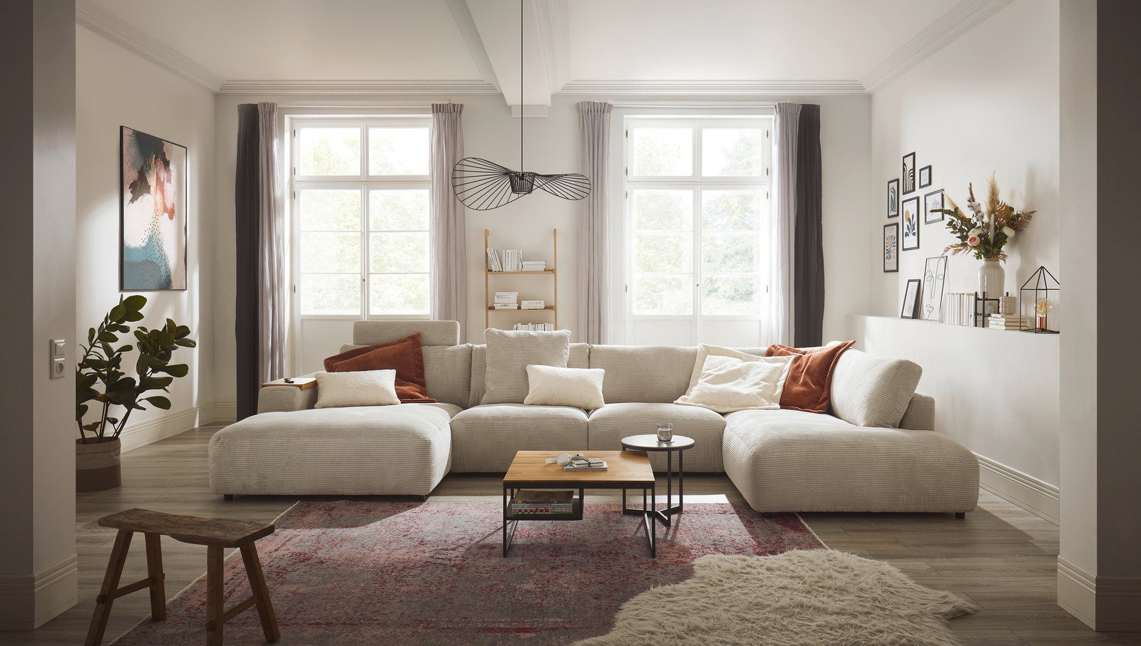 Gemütliches Wohnzimmer mit großem, beigem Ecksofa Musterring LUCIA, dekoriert mit Kissen in Erdtönen, aus der Frontalperspektive fotografiert. Im Hintergrund sind große Fenster, ein Bücherregal und Wanddekorationen zu sehen.