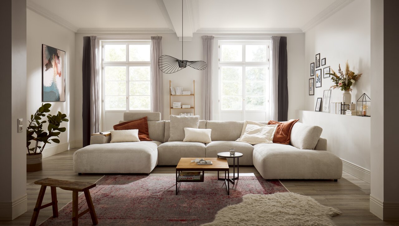 Gezellige woonkamer met een grote, beige hoekbank van het merk Musterring LUCIA, versierd met kussens in aardetinten, gefotografeerd vanuit een vooraanzicht. Op de achtergrond zijn grote ramen, een boekenkast en wanddecoraties te zien.