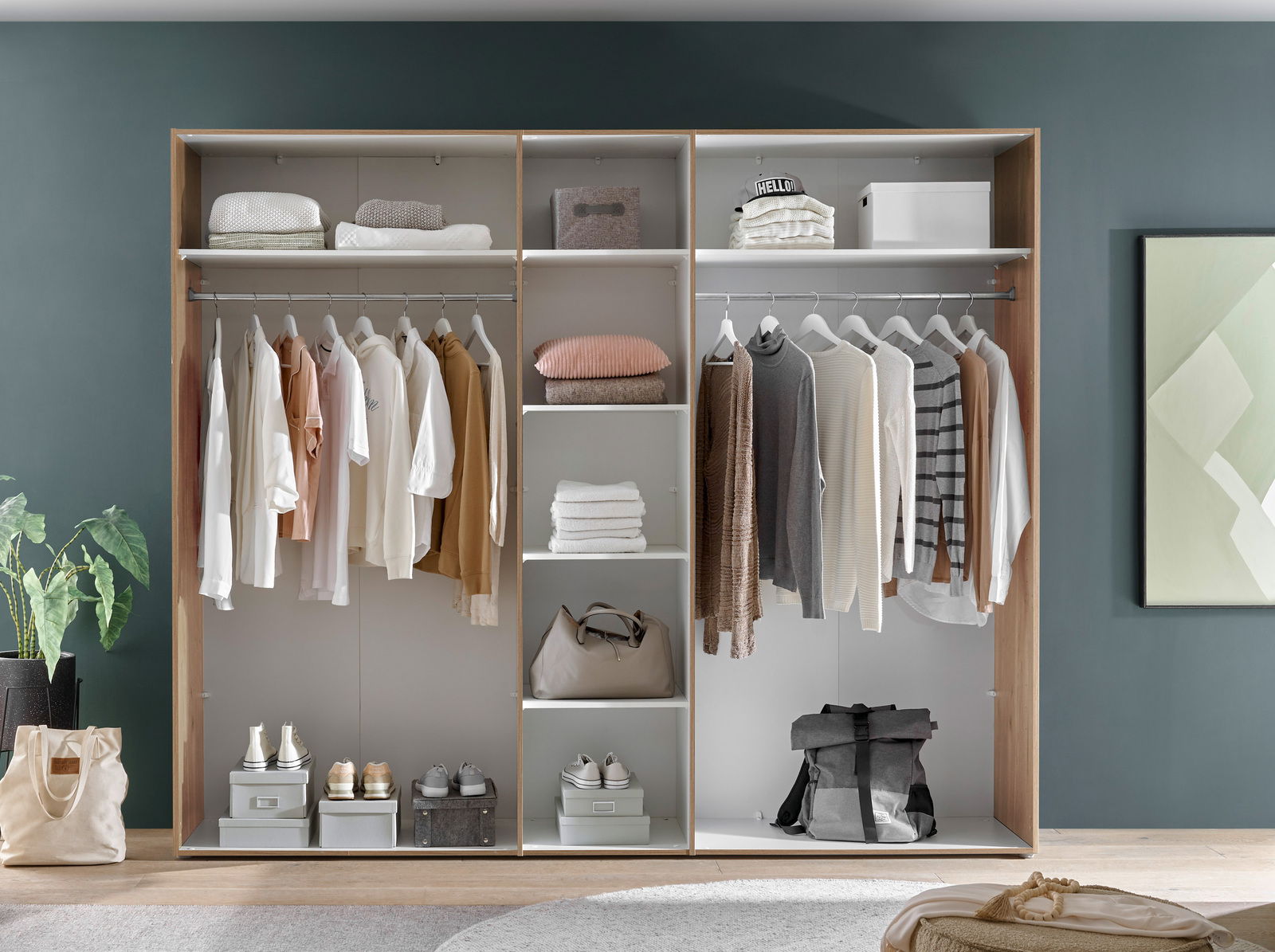 Offener Kleiderschrank mit hängender Kleidung, Regalen mit gefalteten Handtüchern und Taschen, sowie Schuhen auf dem Boden. Frontalansicht.