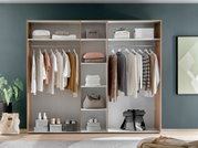 Offener Kleiderschrank mit hängender Kleidung, Regalen mit gefalteten Handtüchern und Taschen, sowie Schuhen auf dem Boden. Frontalansicht.