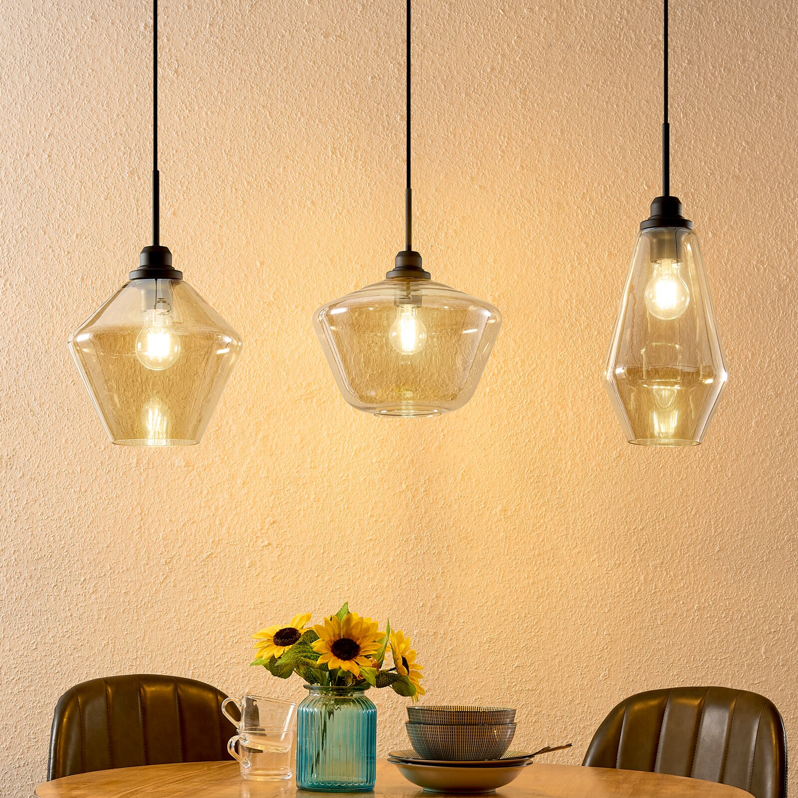 Drie moderne glazen hanglampen in verschillende vormen, die boven een eettafel met bloemen en serviesgoed hangen, gezien vanuit de zijkant.