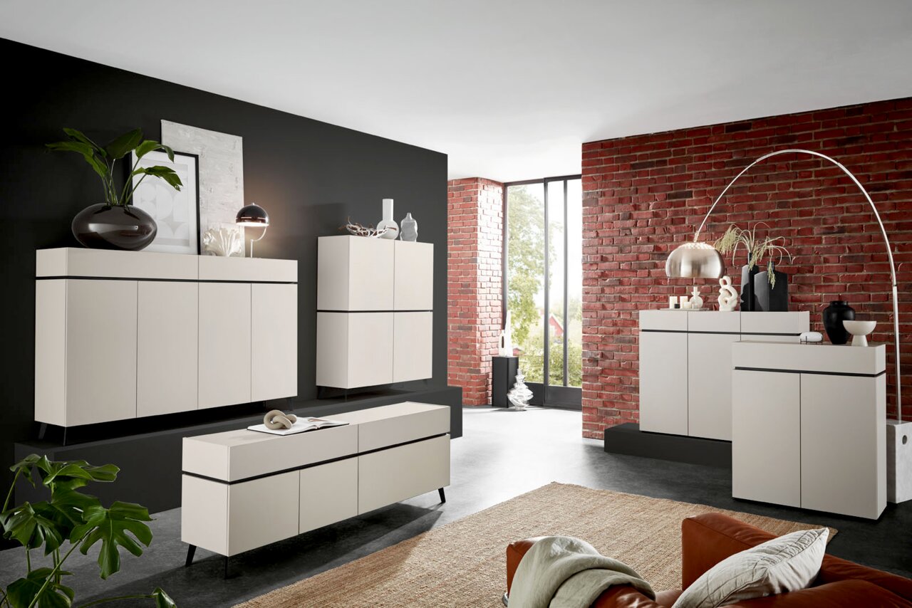 Voleo Fuß-Set UNIQUE Moderne Wohnzimmermöbel in einem stilvollen Raum mit Backsteinwand, bestehend aus Sideboards, Highboards und einem TV-Lowboard in cremefarbenem Design. Perspektive von vorne links.