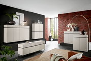 Moderne woonkamermeubels in een stijlvolle ruimte met een bakstenen muur, bestaande uit dressoirs, hoge kasten en een lage tv-kast in een crèmekleurig ontwerp. Aanzicht van linksvoor.