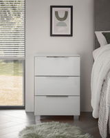 Moderne weiße Nachtkommode mit drei Schubladen, frontal fotografiert, neben einem Bett und einem Fenster mit Jalousien.