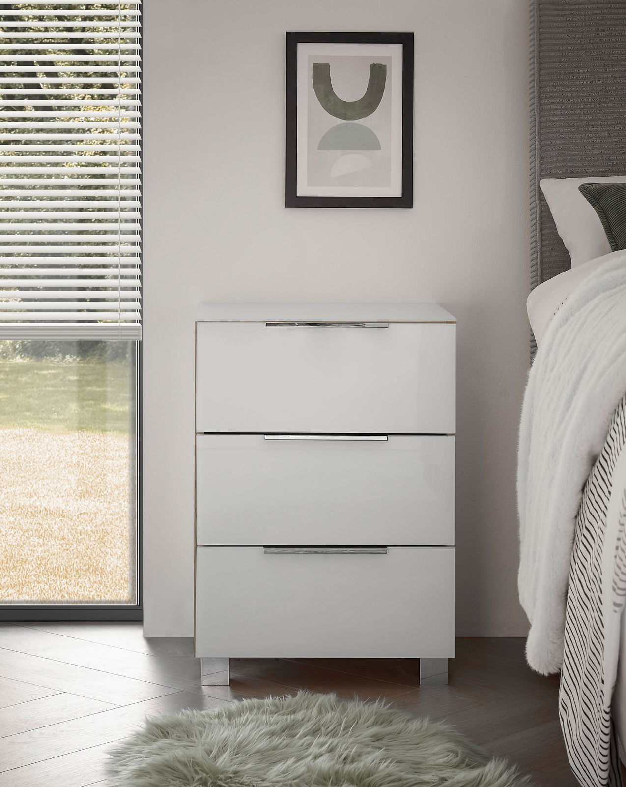 Moderne weiße Nachtkommode mit drei Schubladen, frontal fotografiert, neben einem Bett und einem Fenster mit Jalousien.