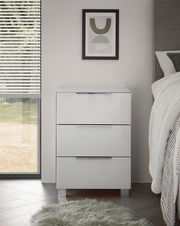Moderne weiße Nachtkommode mit drei Schubladen, frontal fotografiert, neben einem Bett und einem Fenster mit Jalousien.