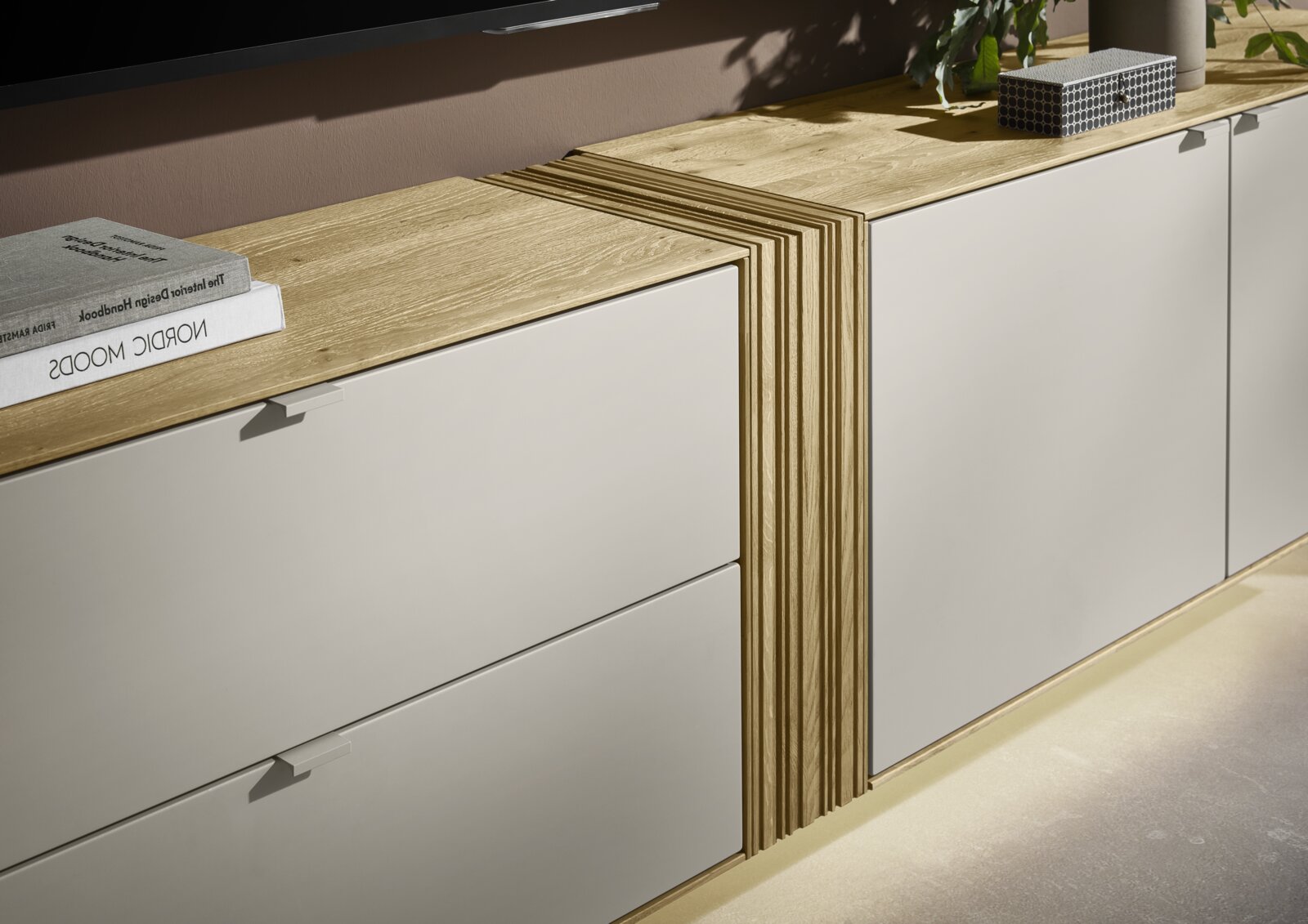 Modernes Lowboard/Sideboard mit hellgrauen Fronten und Holzplatte, mittig geriffeltem Holzelement und aufliegenden Büchern, in schräger Frontansicht von oben.