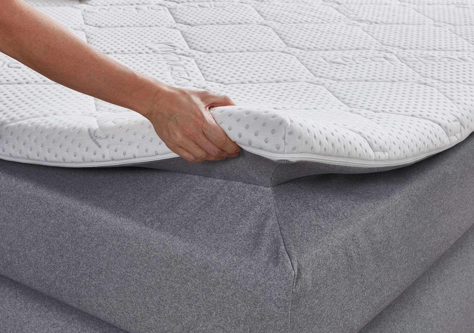 Nahaufnahme eines Boxspringbetts mit einer Hand, die den Matratzentopper anhebt, um die weiche, gepolsterte Oberfläche zu zeigen.